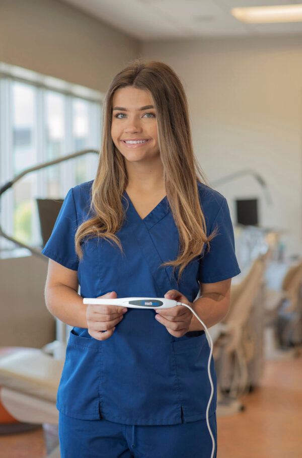 Dental professional in blue scrubs smiling while holding a MouthWatch intraoral camera in a dental office setting.
